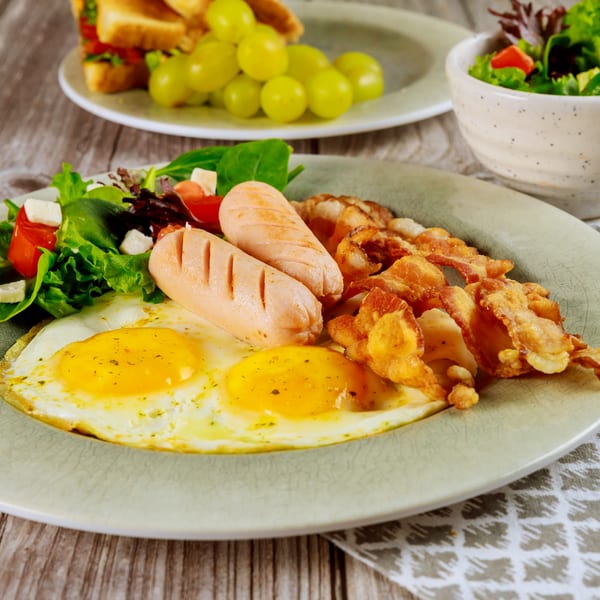 Breakfast plate with fried eggs, sausages, crispy bacon, and a side salad on a wooden table.