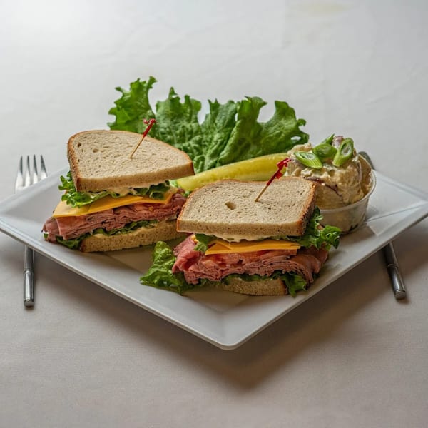 Roast beef and cheese sandwich on whole wheat bread with lettuce, a pickle, and potato salad.