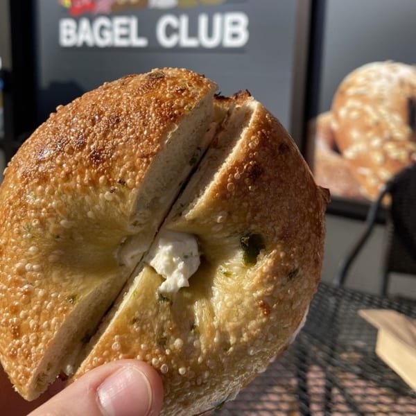 A hand holds a freshly cut bagel with cream cheese, with "BAGEL CLUB" in the background.