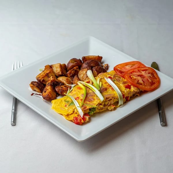 A breakfast plate with a golden omelet topped with scallions, roasted potatoes, and sliced tomatoes.
