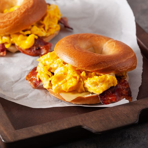 Two Bagel Club breakfast sandwiches with scrambled eggs, bacon, and cheese on a wooden tray.